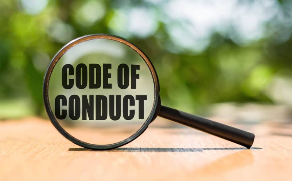 Magnifying glass with text CODE OF CONDUCT on wooden table and green background. Employees of ACS follow this Code of Conduct at their IT Contracting assignments.