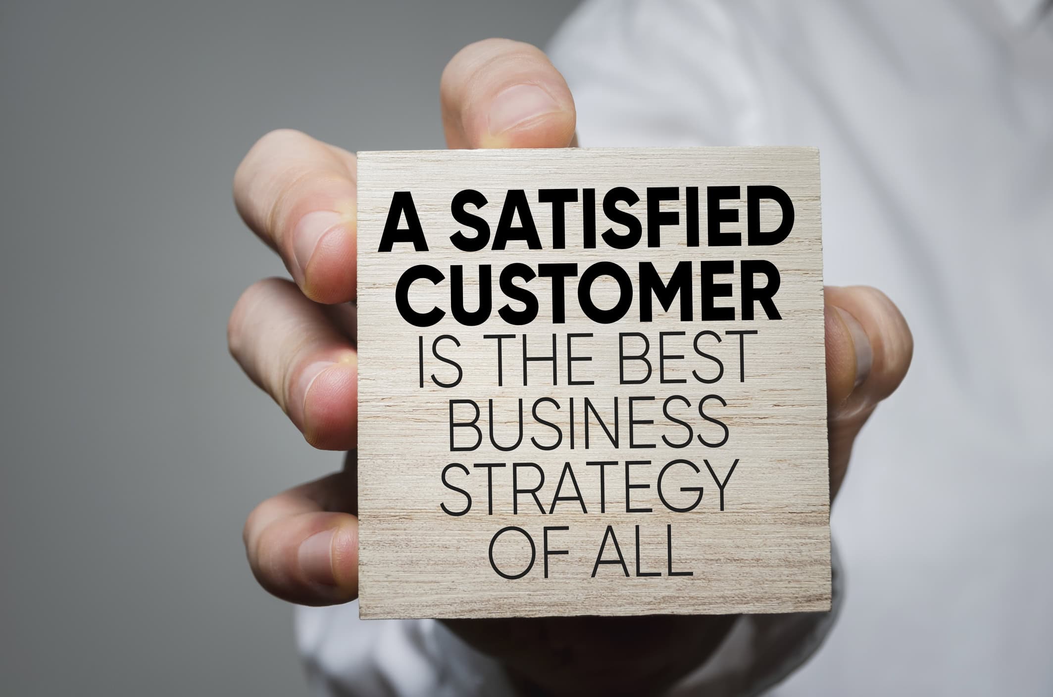 a hand holding a wooden block that has black text saying A Satisfied Customer Is The Best Business Strategy of All indicating that American Computer Services, Inc. delivers satisfaction for Insurance, Technology, and Government Customers.